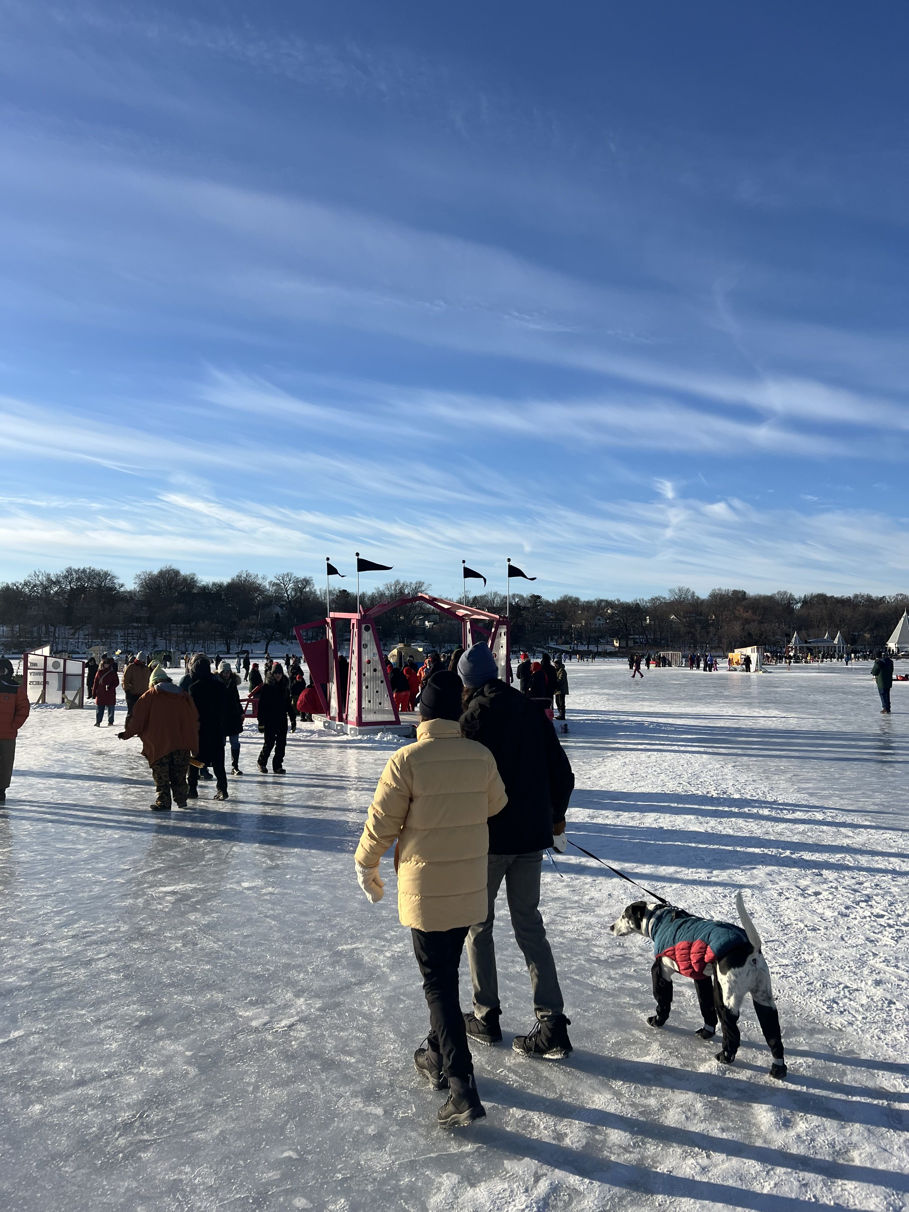 People walking towards art shanty with a dog that has a winter vest and booties on